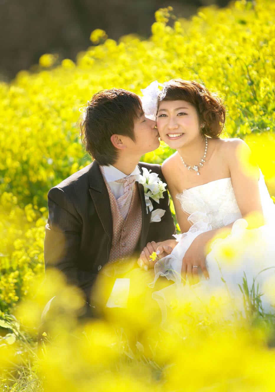 写真：ウェディングドレスを着た新婦とタキシードを着た新郎が菜の花畑でくつろぐ様子